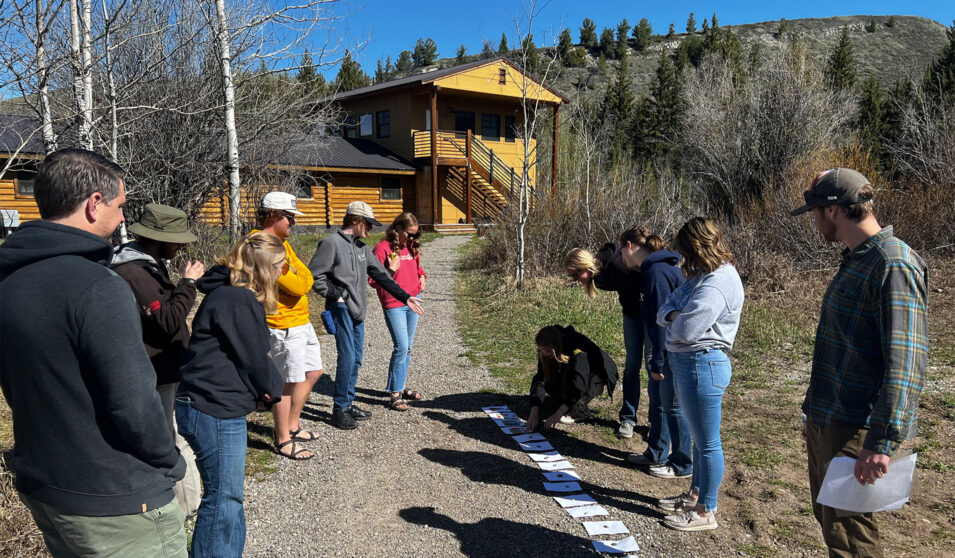 Wyoming Rural Teacher Corps students participating in an activity outside. There are several sheets of white paper on the ground in a row and the students are looking at them. There is a multi-level log cabin like structure behind them and trees along a path leading to the building.