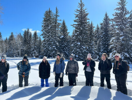 Group of students from Monmouth College in winter gear standing in a semi-circle with snow covered trees behind them.