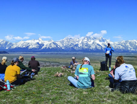 Students from the University of Wyoming Rural Teacher Corps cohort sitting down in the grass with the Teton Mountain range in the background.